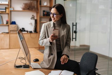 Ofiste otururken cep telefonu ve bilgisayar kullanan gülümseyen kadın.