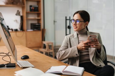 Ofiste bilgisayar ve planlamacıyla çalışırken cep telefonu kullanan sevimli bir kadın.