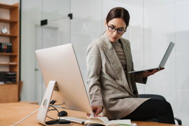 Ofiste bilgisayar ve dizüstü bilgisayarla çalışan gözlüklü çekici bir kadın.