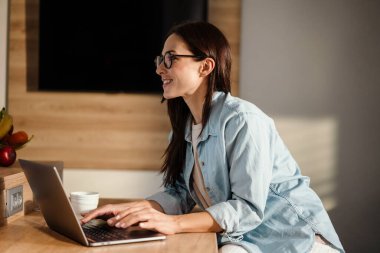 Evde, mutfakta vakit geçirirken dizüstü bilgisayarda çalışan çekici, gülümseyen, zeki bir kadın.