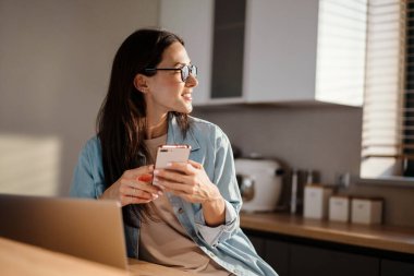 Evde dizüstü bilgisayarla çalışırken akıllı telefon kullanan mutlu bir kadın.