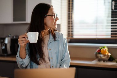 Evde dizüstü bilgisayarla çalışırken kahve içen mutlu bir kadın.