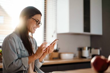 Ev mutfağında akıllı telefon kullanırken gülümseyen mutlu büyüleyici kadın.