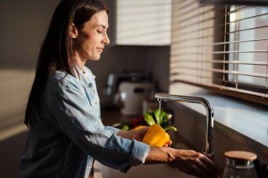 Mutfaktaki lavaboda sebze yıkayan gülümseyen genç kadın.