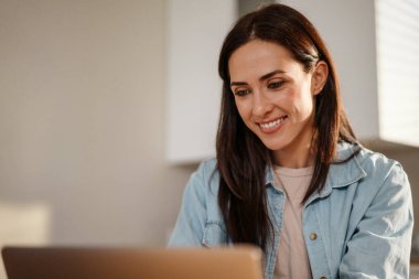 Evde, mutfakta vakit geçirirken dizüstü bilgisayarda çalışan çekici, gülümseyen, zeki bir kadın.