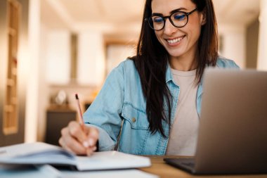 Çekici, gülümseyen, zeki bir kadın dizüstü bilgisayarda çalışırken mutfakta vakit geçirip notlar alıyor.