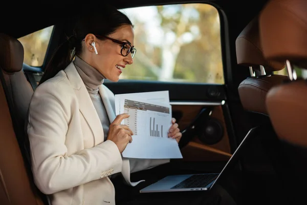 Kulaklıklı Güzel Bir Kadını Arabada Otururken Bilgisayar Kağıtlarla Çalışıyor — Stok fotoğraf