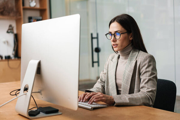 Focused charming woman in eyeglasses working with computer while sitting in office