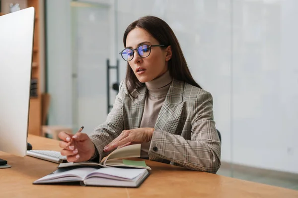 Focused charming woman in eyeglasses working with computer and planner in office