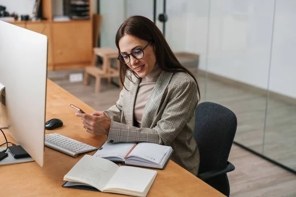 Ofiste bilgisayar ve planlamacıyla çalışırken cep telefonu kullanan hoş bir kadın.