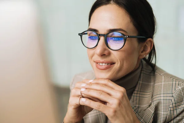 Smiling charming woman in eyeglasses working with computer while sitting in office