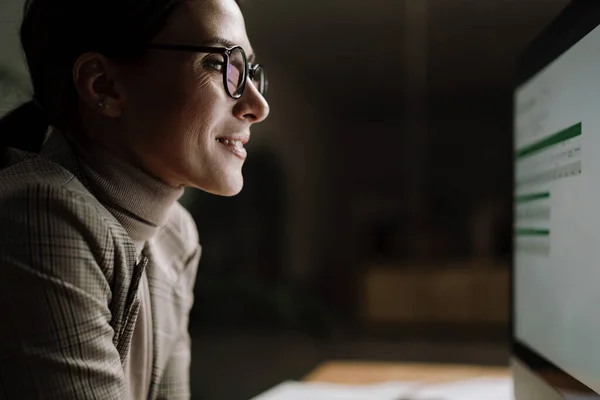 オフィスに座っている間にコンピュータで働く眼鏡の中で魅力的な女性を笑顔 — ストック写真