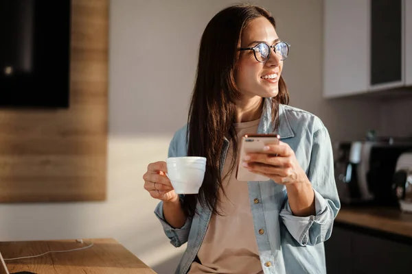 自宅のキッチンでスマートフォンやコーヒーを飲んで幸せな魅力的な女性 — ストック写真