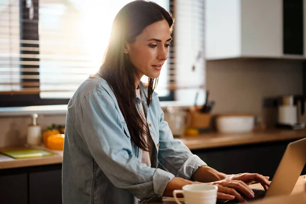 Attractive smiling young smart woman working on laptop computer while spending time at home in the kitchen