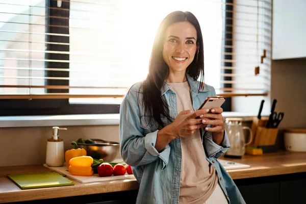 笑顔魅力的な若い女性テキストメッセージの携帯電話でザキッチン — ストック写真