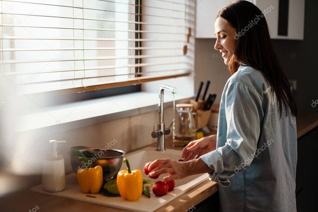 Smiling young woman washing vegetable in the sink in kitchen