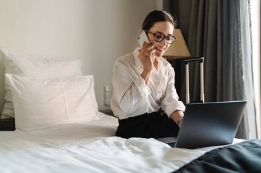 Orta yaşlı, esmer, beyaz bir iş kadını otel odasında oturmuş cep telefonuyla dizüstü bilgisayar kullanarak konuşuyor.