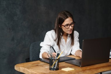 Düşünceli, orta yaşlı esmer kadın, karanlık duvar arka planında dizüstü bilgisayarda çalışıyor.