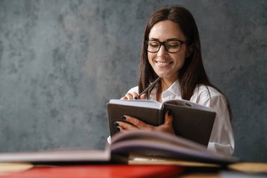 Orta yaşlı, esmer, gülümseyen bir kadın. Masada oturmuş, arka planda koyu renk duvar kitapları okuyor.