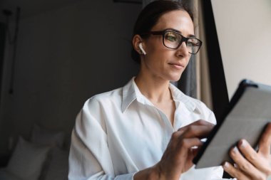 Gülümseyen orta yaşlı esmer, akıllı beyaz iş kadını otel odasının penceresinde kablosuz kulaklık takıp tablet cihazına dokunuyor.