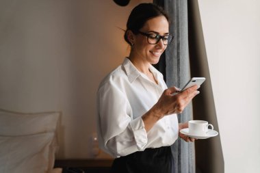 Gülümseyen orta yaşlı esmer beyaz iş kadını. Otel odasının penceresinde elinde bir fincan çayla cep telefonuna bakıyor.