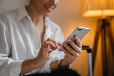 Gülümseyen orta yaşlı esmer beyaz iş kadını otel odasında oturmuş cep telefonuyla mesaj atıyor.