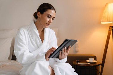 Gülümseyen orta yaşlı esmer, bornozlu beyaz kadın elinde tablet bilgisayarla yatağında oturuyor.