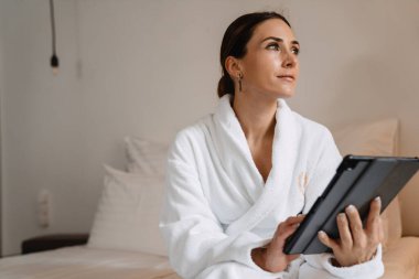 Gülümseyen orta yaşlı esmer, bornozlu beyaz kadın elinde tablet bilgisayarla yatağında oturuyor.