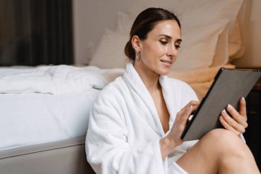 Gülümseyen orta yaşlı esmer, bornozlu beyaz kadın, elinde tablet bilgisayarla bir yatakta oturuyor.