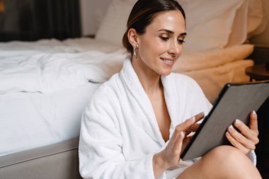 Gülümseyen orta yaşlı esmer, bornozlu beyaz kadın, elinde tablet bilgisayarla bir yatakta oturuyor.