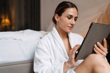 Gülümseyen orta yaşlı esmer, bornozlu beyaz kadın, elinde tablet bilgisayarla bir yatakta oturuyor.