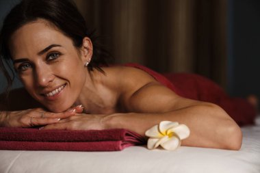 Orta yaşlı esmer bir kadın havluya sarılı bir masanın üzerine uzanmış, spa salonunda gülümsüyor.