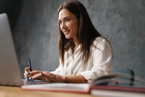 穿着衬衫的中年黑发女商人坐在办公桌前 带着笔记本电脑在黑暗的背景下微笑 — 图库照片