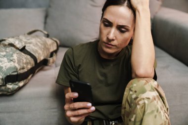 Kapalı alanda otururken cep telefonu kullanan ciddi ve güzel bir kadın asker.