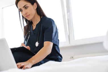 Evde dizüstü bilgisayarla çalışırken steteskopla yazan odaklanmış kadın doktor.