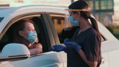 Koruyucu yüz maskesi takan bir kadın işçi arabada oturan bir kadın sürücüyle konuşuyor.