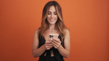 Rüya gören yetişkin bir kadın, stüdyoda turuncu arka planda izole bir şekilde cep telefonunu kullanıyor.