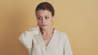 Kafası karışmış orta yaşlı esmer bir kadın omuzlarını silkiyor. Stüdyoda bej arka planda tek başına duruyor.
