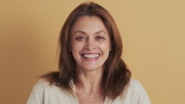 Mutlu orta yaşlı esmer bir kadın gülerek stüdyoda tek başına bej arka planda duruyor.