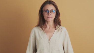 Kızgın, orta yaşlı, gözlüklü bir esmer kadın stüdyoda izole edilmiş bej arkaplanda kameraya bakıyor.