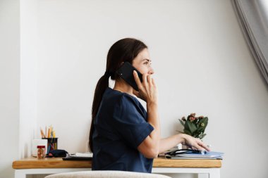 Masada oturan ve telefonda konuşan kadın doktorun yan görüntüsü.