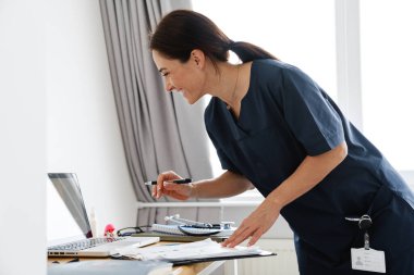 Gülümseyen bir doktor dizüstü bilgisayarın önünde duruyor ve parlak bir odada video konuşmalarıyla konuşuyor.