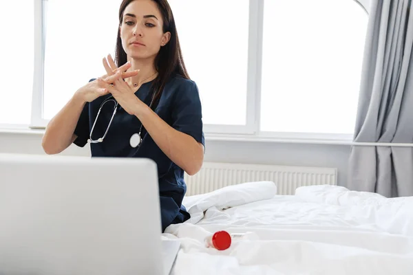 Brunette woman doctor in a medical suit showing how to properly wash her hands by video call, sitting on the bed against the background of a window