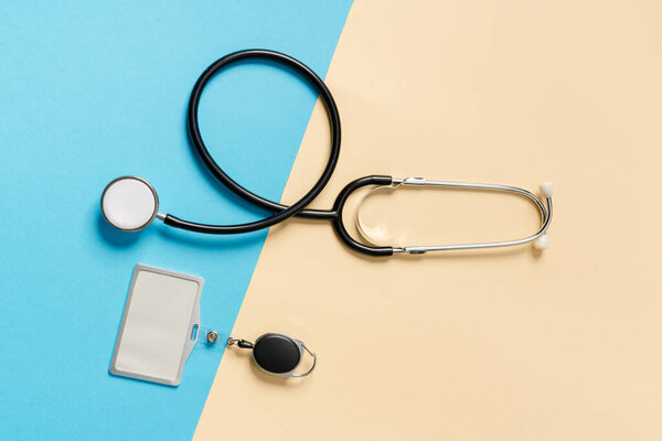 Overhead view of a stethoscope with a badge lying sideways on a beige and blue background in the studio