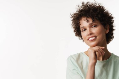 Genç kıvırcık esmer kadın gülümsüyor ve beyaz arka planda izole edilmiş kameraya bakıyor.