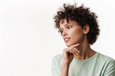 Genç kıvırcık esmer kadın beyaz arka planda yalnız poz veriyor ve yan bakıyor.