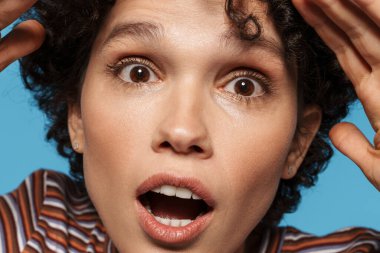 Genç kıvırcık kadın mavi arka planda mahsur kalmış kameraya şaşırdığını ifade ediyor.