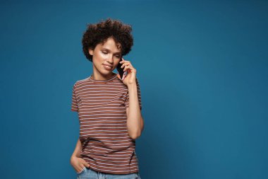 Çizgili tişörtlü genç, kıvırcık kadın mavi arka planda cep telefonuyla konuşuyor.