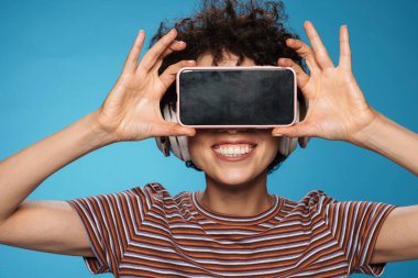 Genç, kıvırcık, gülümseyen, kulaklık takan cep telefonlu bir kadın.