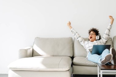 Kulaklıklı genç bir kadın evde kanepede otururken esniyor ve dizüstü bilgisayar kullanıyor.